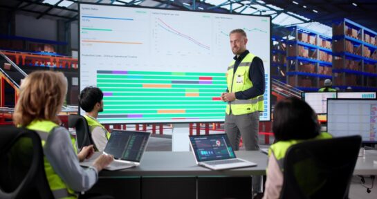 Team aziendale analizza dati commerciali su un grande schermo all’interno di uno stabilimento industriale